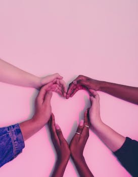 A group of diverse hands forming a heart shape against a soft pink background, symbolizing love and unity.
